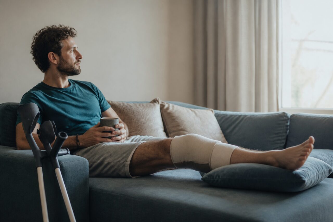 Personne au repos sur un canapé, la jambe opérée surélevée sur un coussin, béquilles à proximité